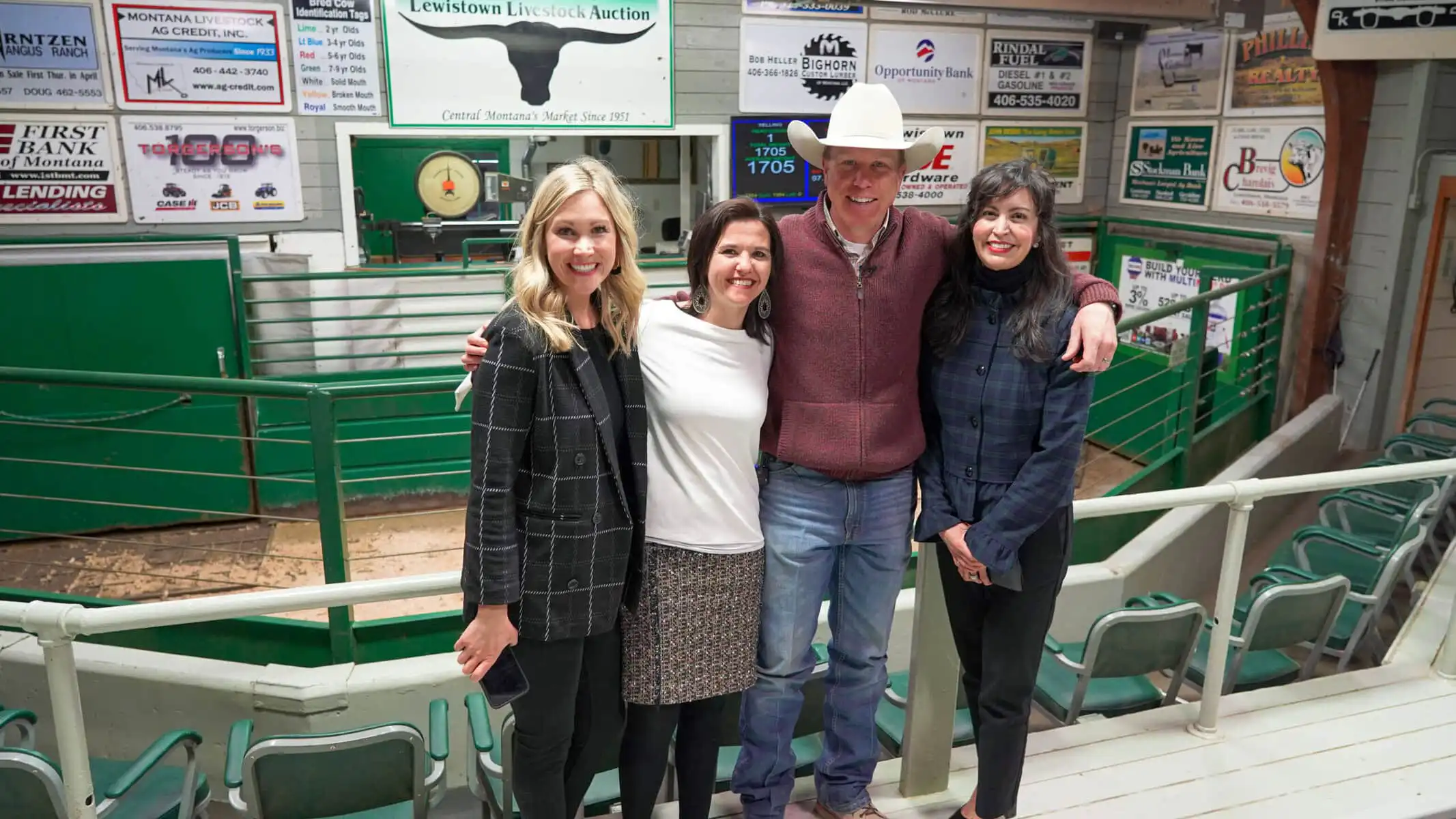 Four people stand smiling with arms around each other inside the Lewistown indoor livestock auction arena, with green fencing and advertisements on the walls in the background.