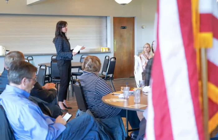 A woman stands and speaks to a seated group in a Lewistown meeting room. People listen attentively, some with papers and drinks on the tables. An American flag is partially visible as election officials gather for Secretary Jacobsens update.