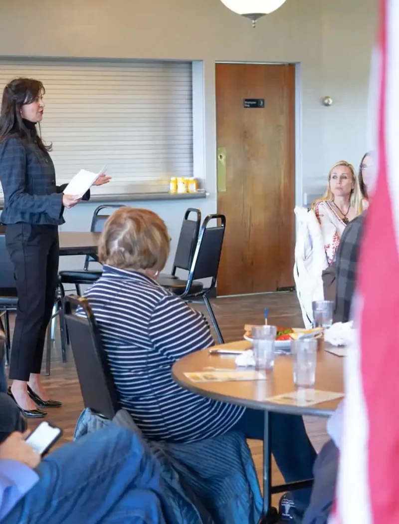 Secretary Jacobsen visits Lewistown; meets election officials, business owners, and students 7 A woman stands and speaks to a seated group in a Lewistown meeting room. People listen attentively, some with papers and drinks on the tables. An American flag is partially visible as election officials gather for Secretary Jacobsens update.