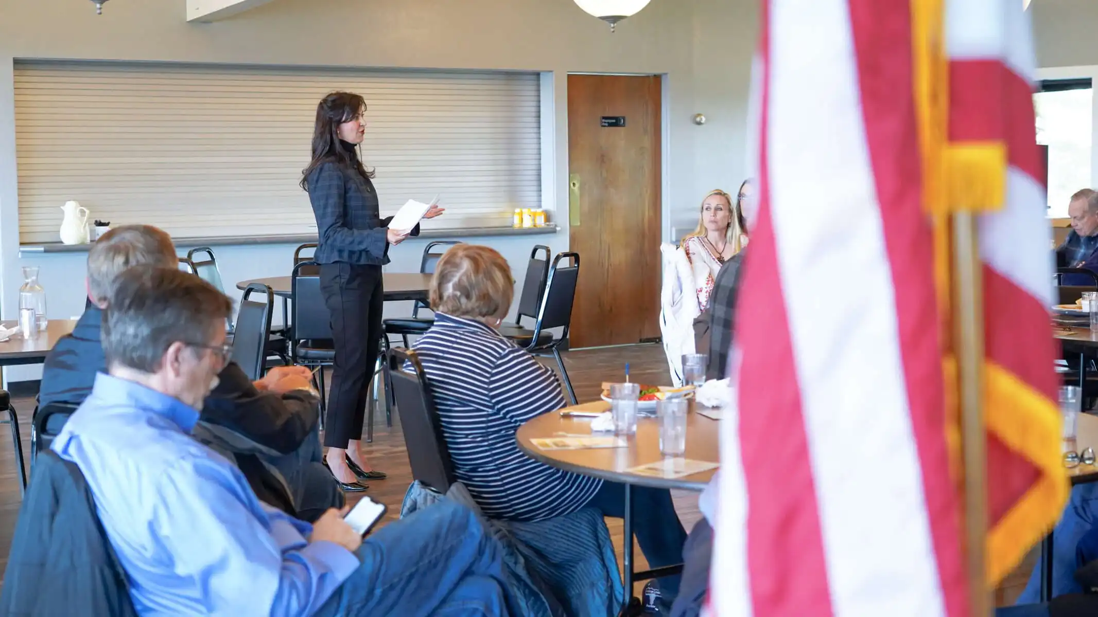 A woman stands and speaks to a seated group in a Lewistown meeting room. People listen attentively, some with papers and drinks on the tables. An American flag is partially visible as election officials gather for Secretary Jacobsens update.