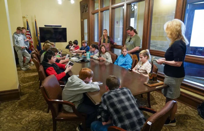 Corvallis 7th-graders meet with Secretary of State's Office as part of field trip 4 Corvallis 7th-graders sit around a large conference table in a formal room during their field trip to the Secretary of States Office. Some use devices as several adults stand nearby, with two American flags visible in the background.