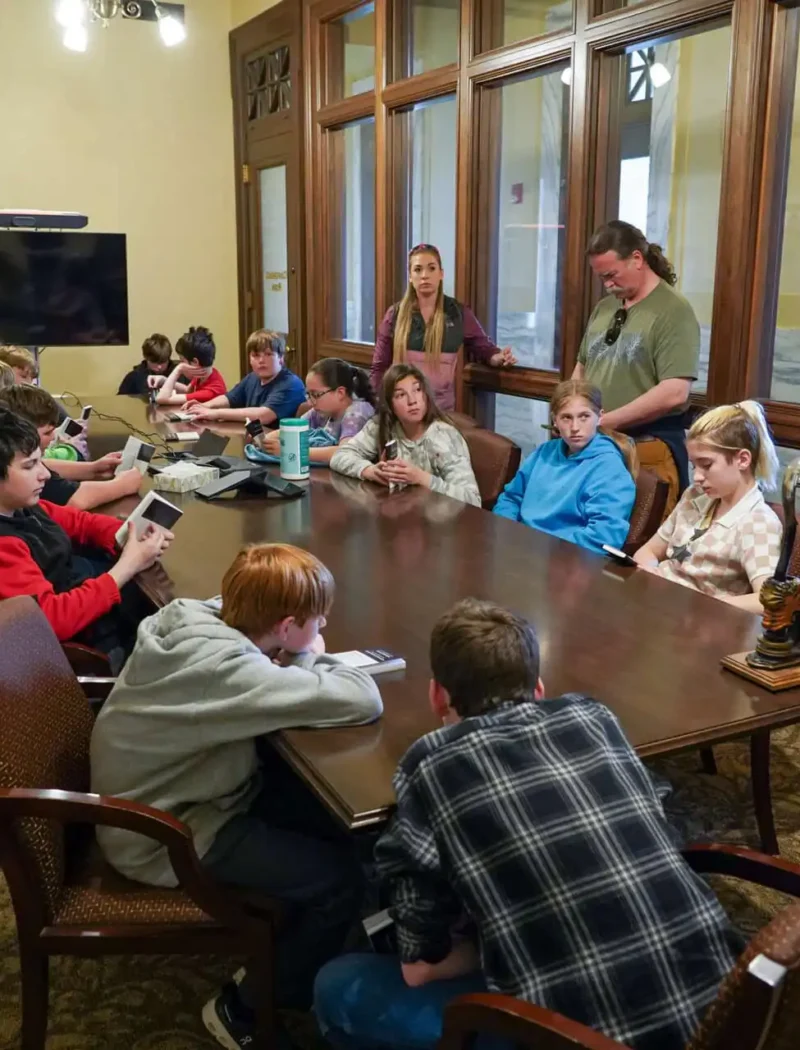 Corvallis 7th-graders sit around a large conference table in a formal room during their field trip to the Secretary of States Office. Some use devices as several adults stand nearby, with two American flags visible in the background.
