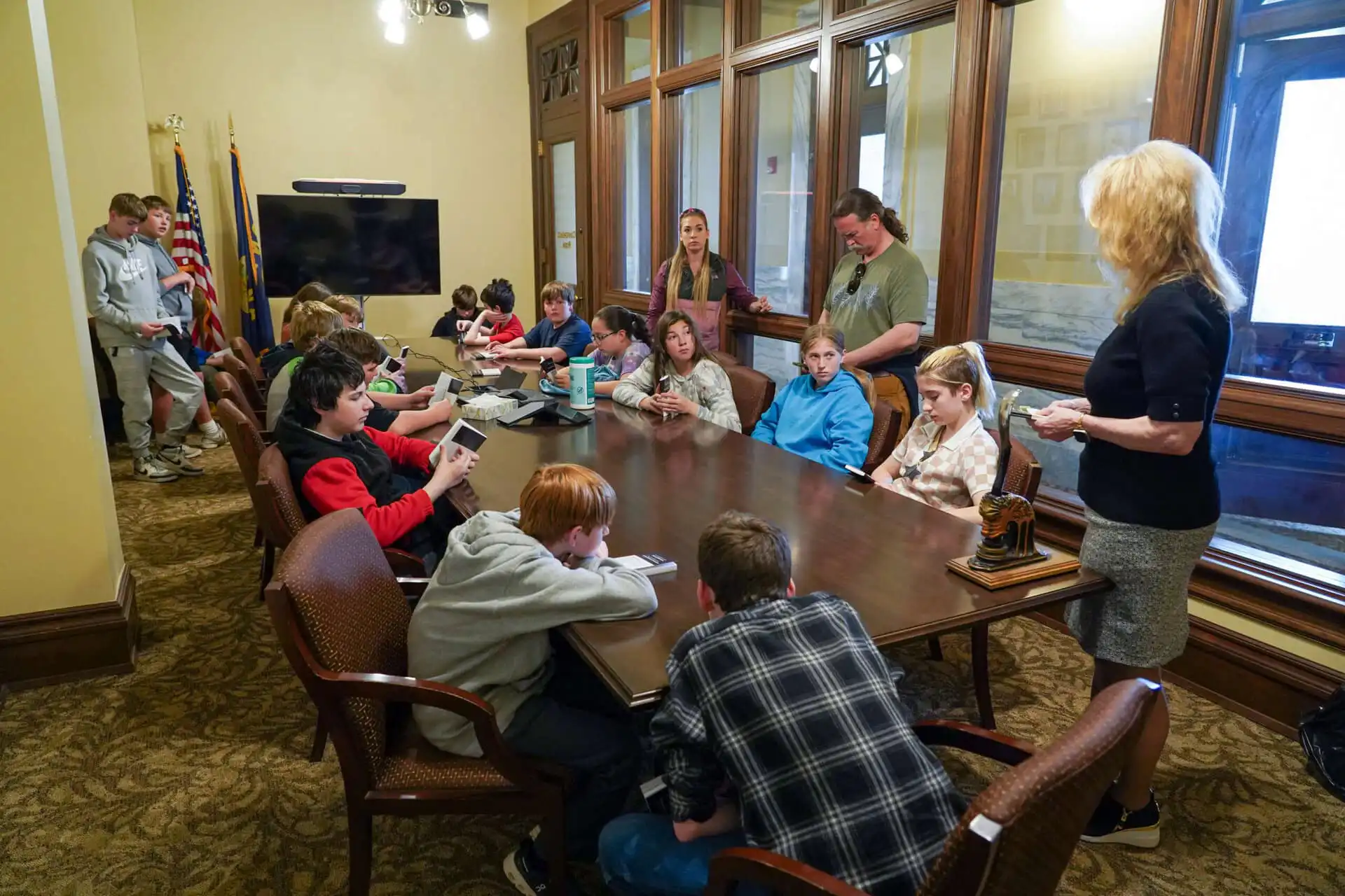 Corvallis 7th-graders sit around a large conference table in a formal room during their field trip to the Secretary of States Office. Some use devices as several adults stand nearby, with two American flags visible in the background.