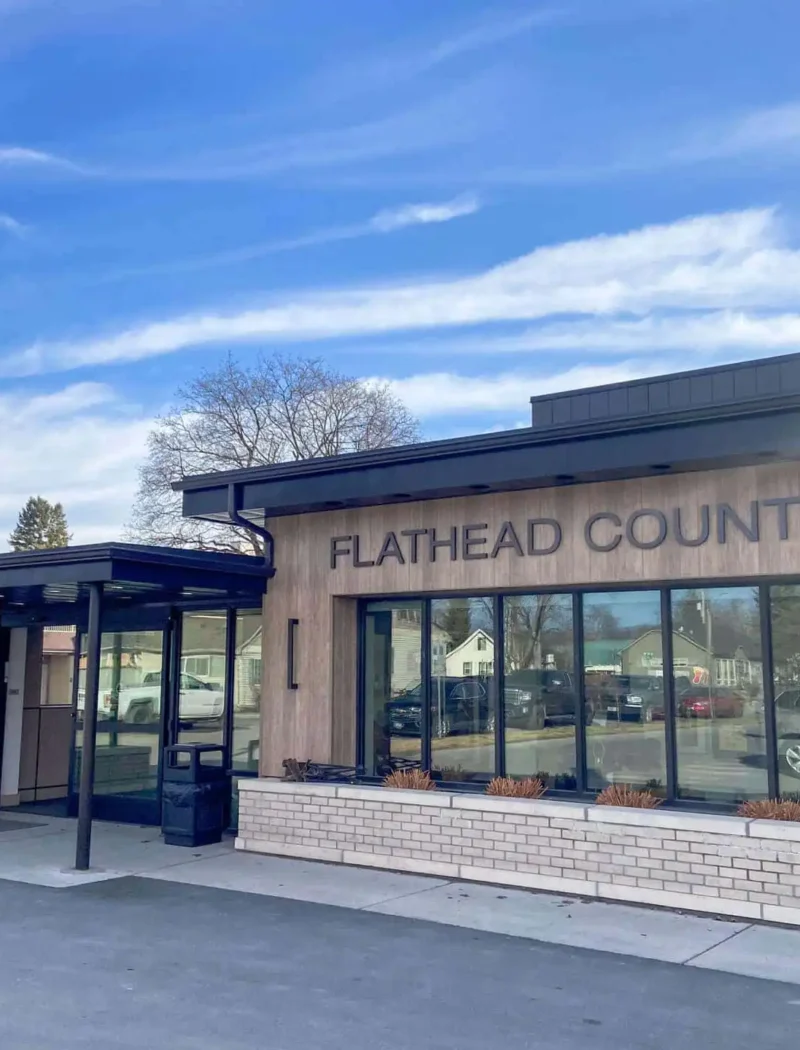 A beige building with large windows displays the words FLATHEAD COUNTY on its exterior under a blue sky with wispy clouds. An American flag stands near the entrance, highlighting where Flathead County elections are held.