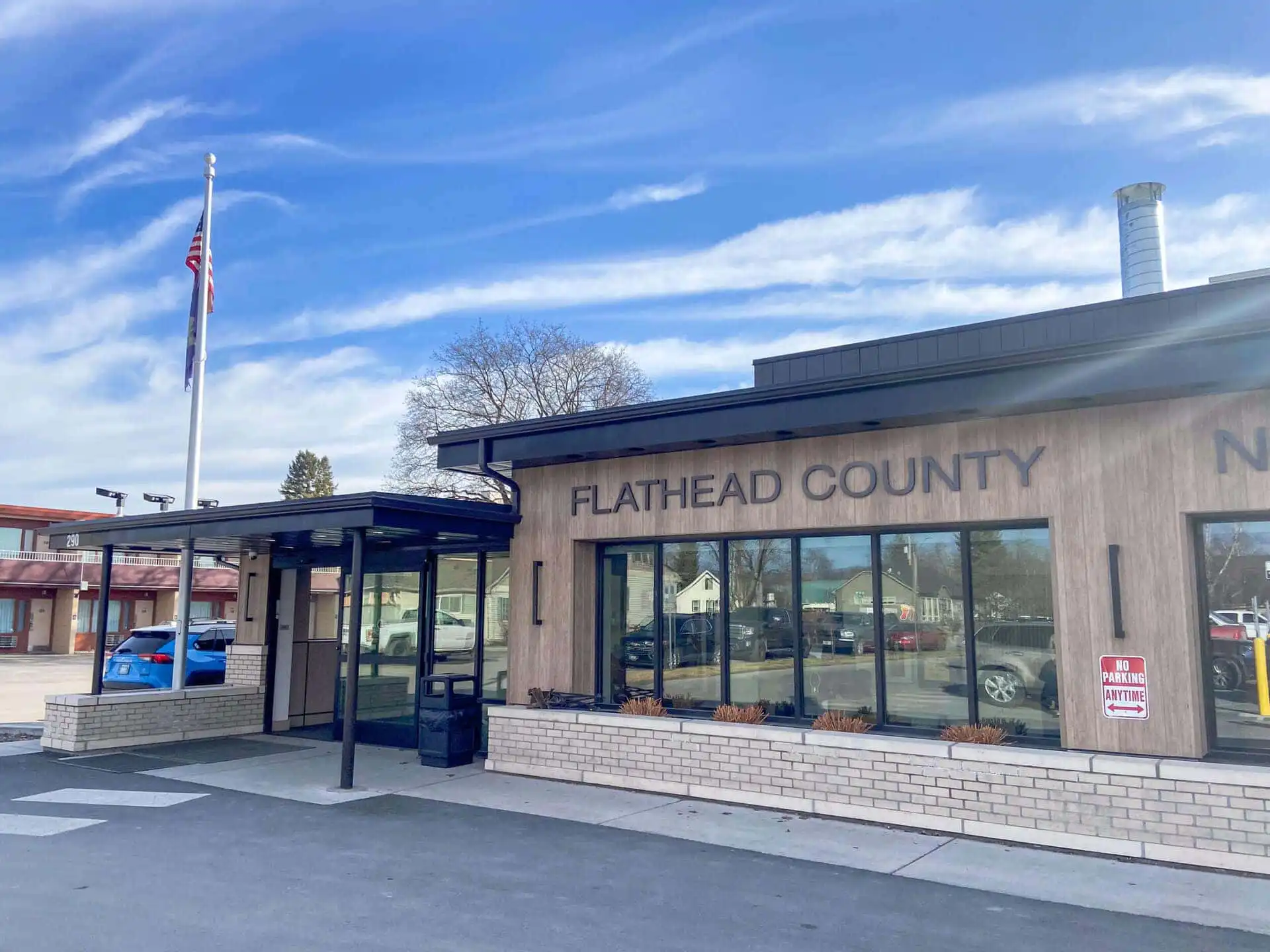 A beige building with large windows displays the words FLATHEAD COUNTY on its exterior under a blue sky with wispy clouds. An American flag stands near the entrance, highlighting where Flathead County elections are held.
