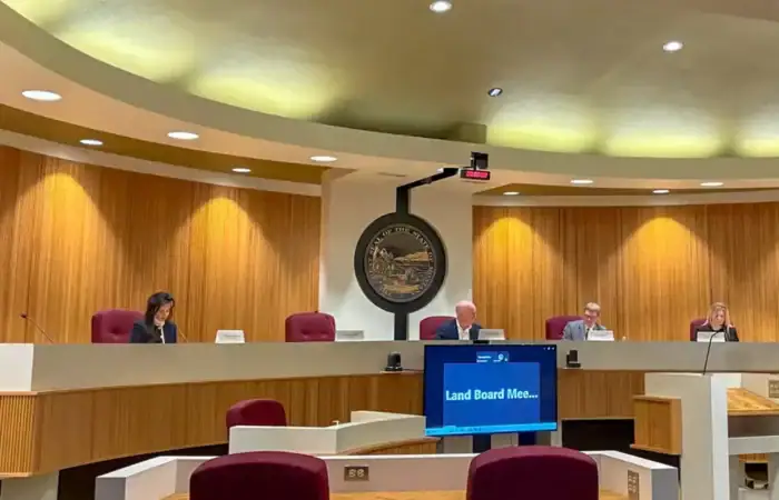 A government meeting room with four people seated at a curved desk, microphones in front of them. A large seal is on the wall, and a screen reads Land Board Mee...—Christi Jacobsen discusses a logging project in Lincoln County. Empty red chairs sit in the foreground.
