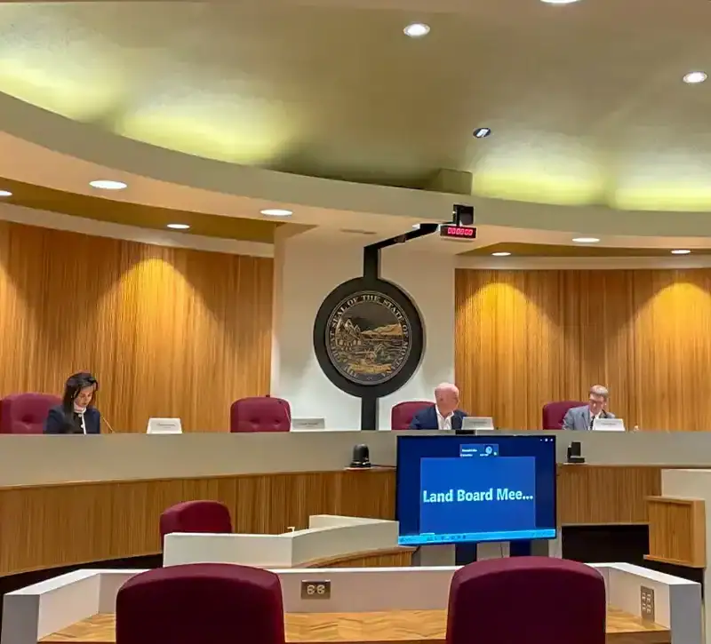 A government meeting room with four people seated at a curved desk, microphones in front of them. A large seal is on the wall, and a screen reads Land Board Mee...—Christi Jacobsen discusses a logging project in Lincoln County. Empty red chairs sit in the foreground.