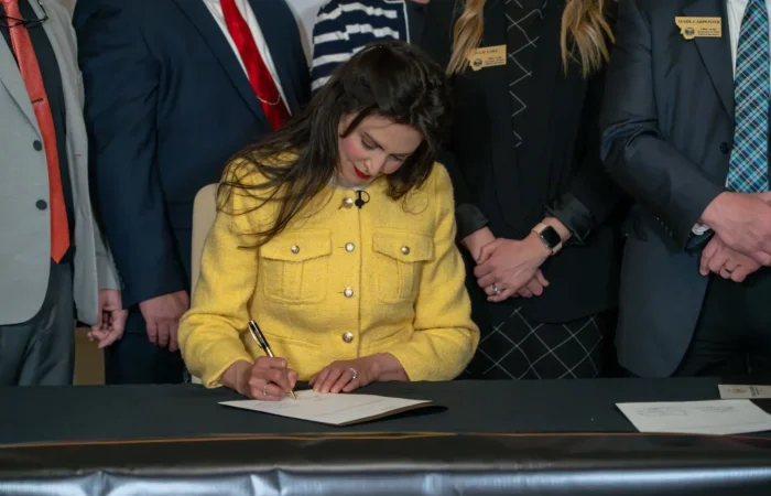 Secretary Christi Jacobsen: 'Bill to enhance Montana's voter registration opportunities heads to Governor's desk' 6 A woman in a yellow jacket sits at a table, signing a document for the Death of a Loved One Initiative, surrounded by several people—including Christi Jacobsen and Montana funeral directors—who stand behind her in formal attire.