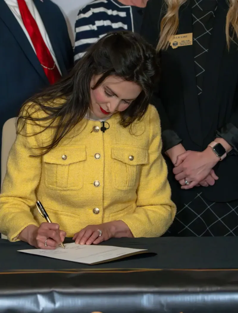 A woman in a yellow jacket sits at a table, signing a document for the Death of a Loved One Initiative, surrounded by several people—including Christi Jacobsen and Montana funeral directors—who stand behind her in formal attire.