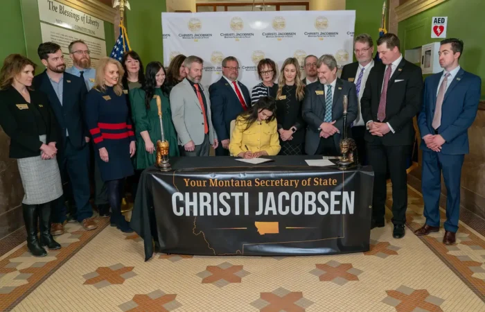 Secretary Christi Jacobsen, Montana funeral directors celebrate 'Death of a Loved One Initiative' being signed into law 8 A woman sits and signs a document at a table labeled “Your Montana Secretary of State Christi Jacobsen,” surrounded by a group of people, with an American flag and official banners in the background.