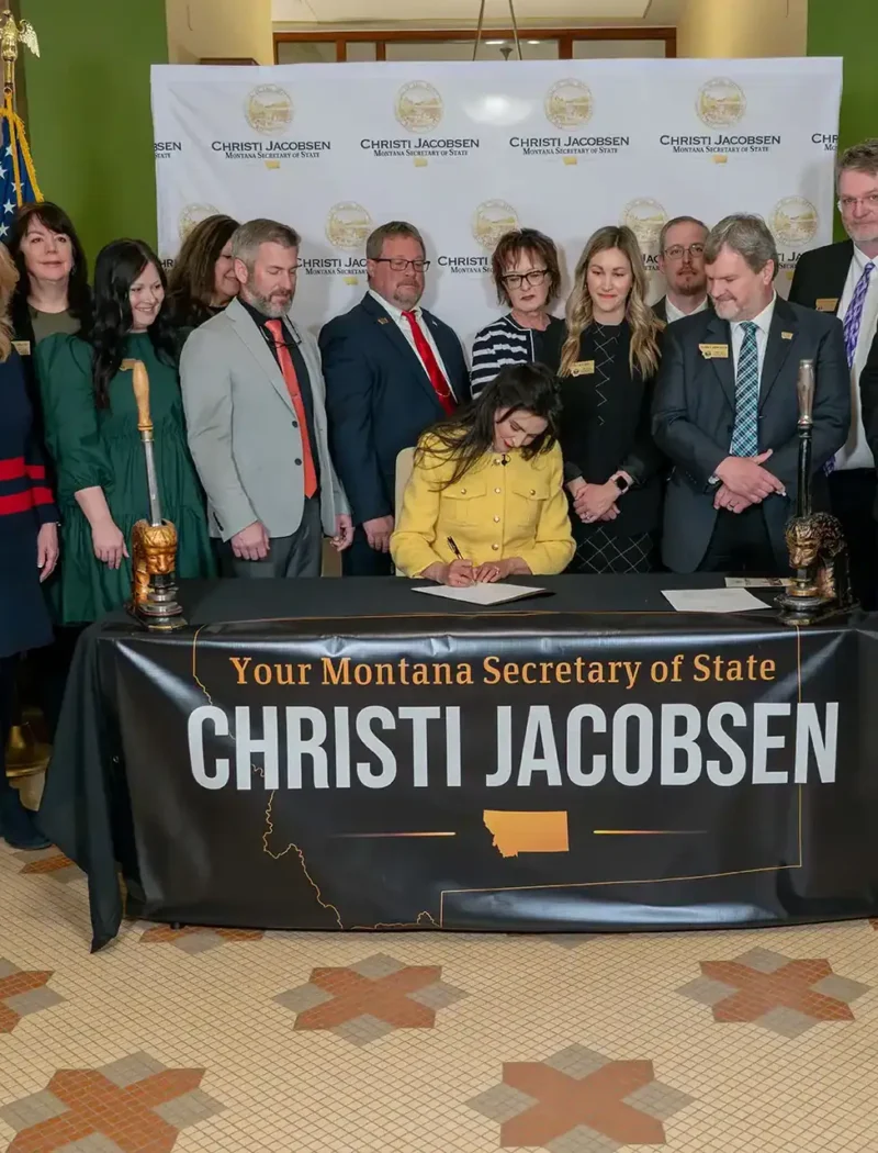 A woman sits and signs a document at a table labeled “Your Montana Secretary of State Christi Jacobsen,” surrounded by a group of people, with an American flag and official banners in the background.