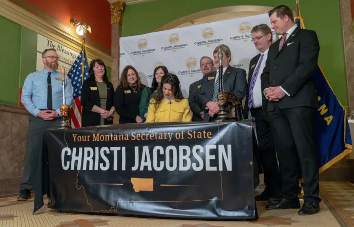 Secretary Christi Jacobsen: 'Montana’s photo voter ID bill heads to Governor's desk' 7 A group of people, some standing and one seated, gather around a desk—possibly the Governors desk—with a banner reading “Your Montana Secretary of State Christi Jacobsen.” U.S. and Montana flags are in the background; the setting appears official.