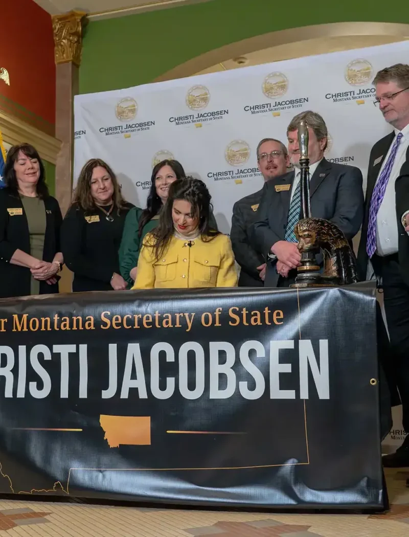 A group of people, some standing and one seated, gather around a desk—possibly the Governors desk—with a banner reading “Your Montana Secretary of State Christi Jacobsen.” U.S. and Montana flags are in the background; the setting appears official.