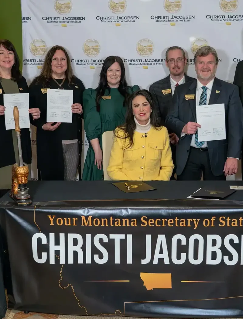 A group of people stand behind a seated woman at a table with a sign reading Your Montana Secretary of State Christi Jacobsen. Celebrating red tape relief, several people hold documents and smile at the camera.