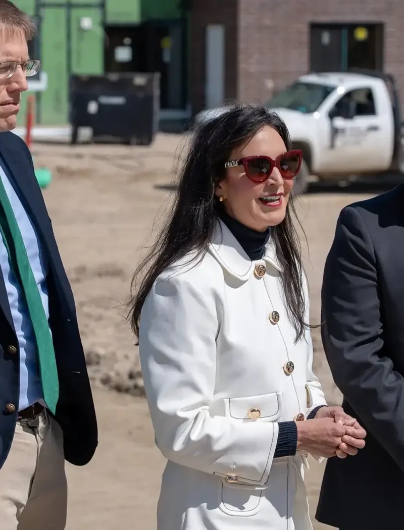 Secretary Christi Jacobsen stands at a construction site in April 2025 with two colleagues, highlighting the surge in new business registrations. The woman wears a white coat and sunglasses, flanked by two men in suits, with vehicles and materials in the background.