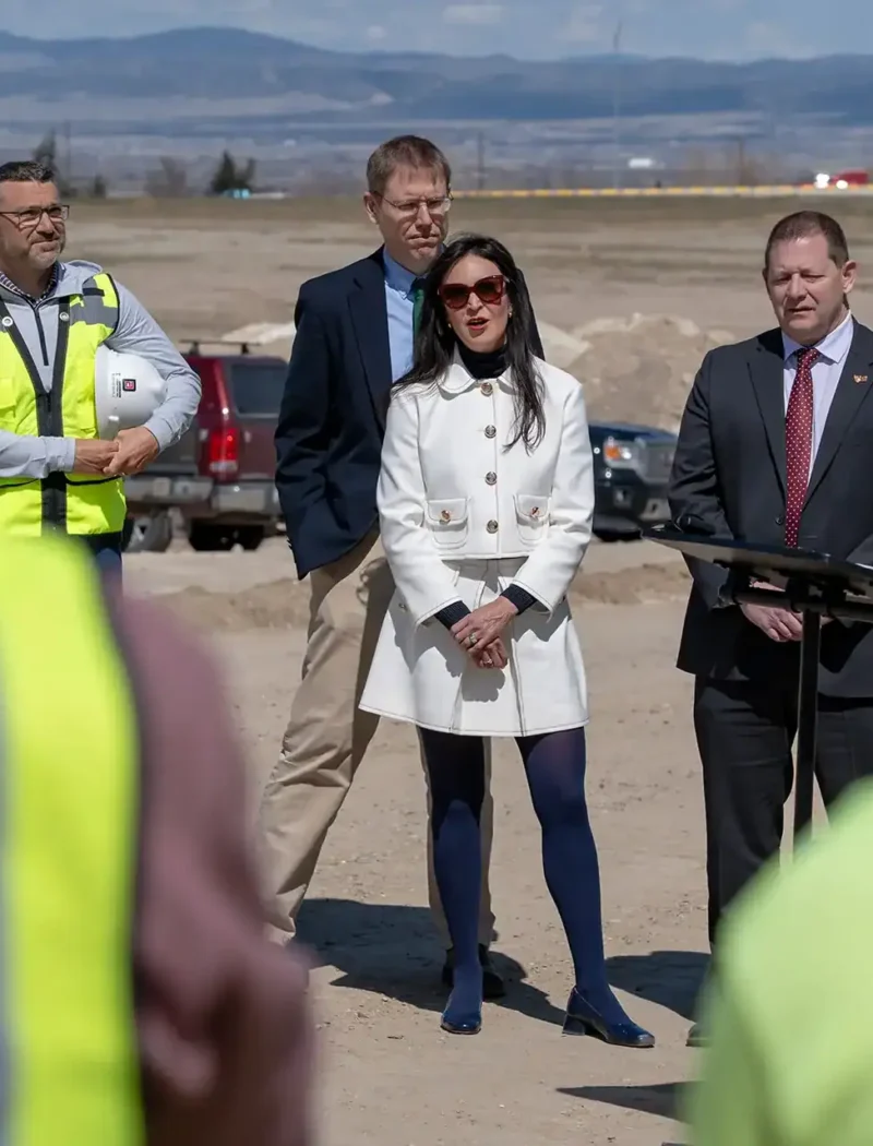 Secretary Christi Jacobsen addresses the crowd at a podium outdoors, flanked by officials. People in high-visibility vests stand nearby, while construction vehicles and Montana businesses are set against a mountainous backdrop.