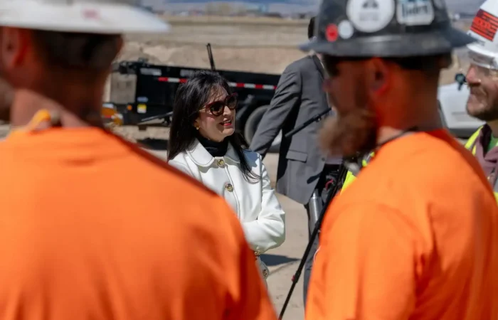 Secretary Christi Jacobsen: 'Montana's pro-business environment fuels 6,500 new business registrations in May 2025 3 A woman in a white coat and sunglasses stands outdoors, talking to two construction workers in orange shirts and hard hats—reflecting Montana’s pro-business environment. Construction equipment and a person in a suit are visible in the background.