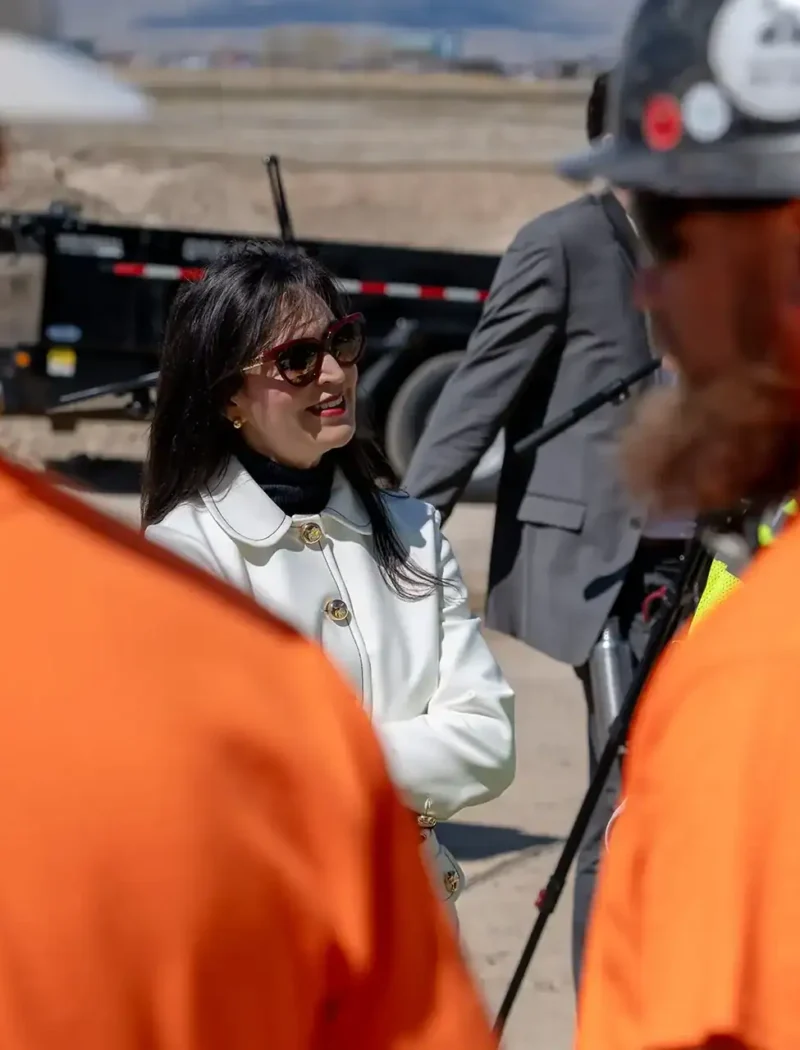 A woman in a white coat and sunglasses stands outdoors, talking to two construction workers in orange shirts and hard hats—reflecting Montana’s pro-business environment. Construction equipment and a person in a suit are visible in the background.