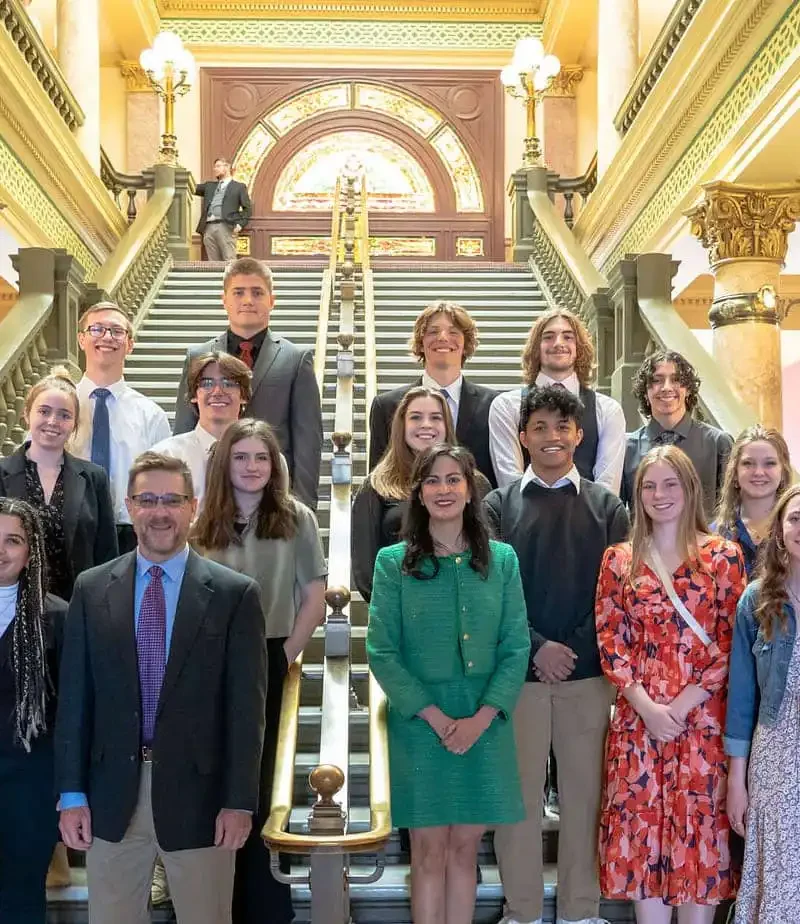 A group of people, including students from Stevensville High School and several adults, stand posing for a photo on a grand staircase inside a historic building with ornate columns, decorative lights, and colorful walls.