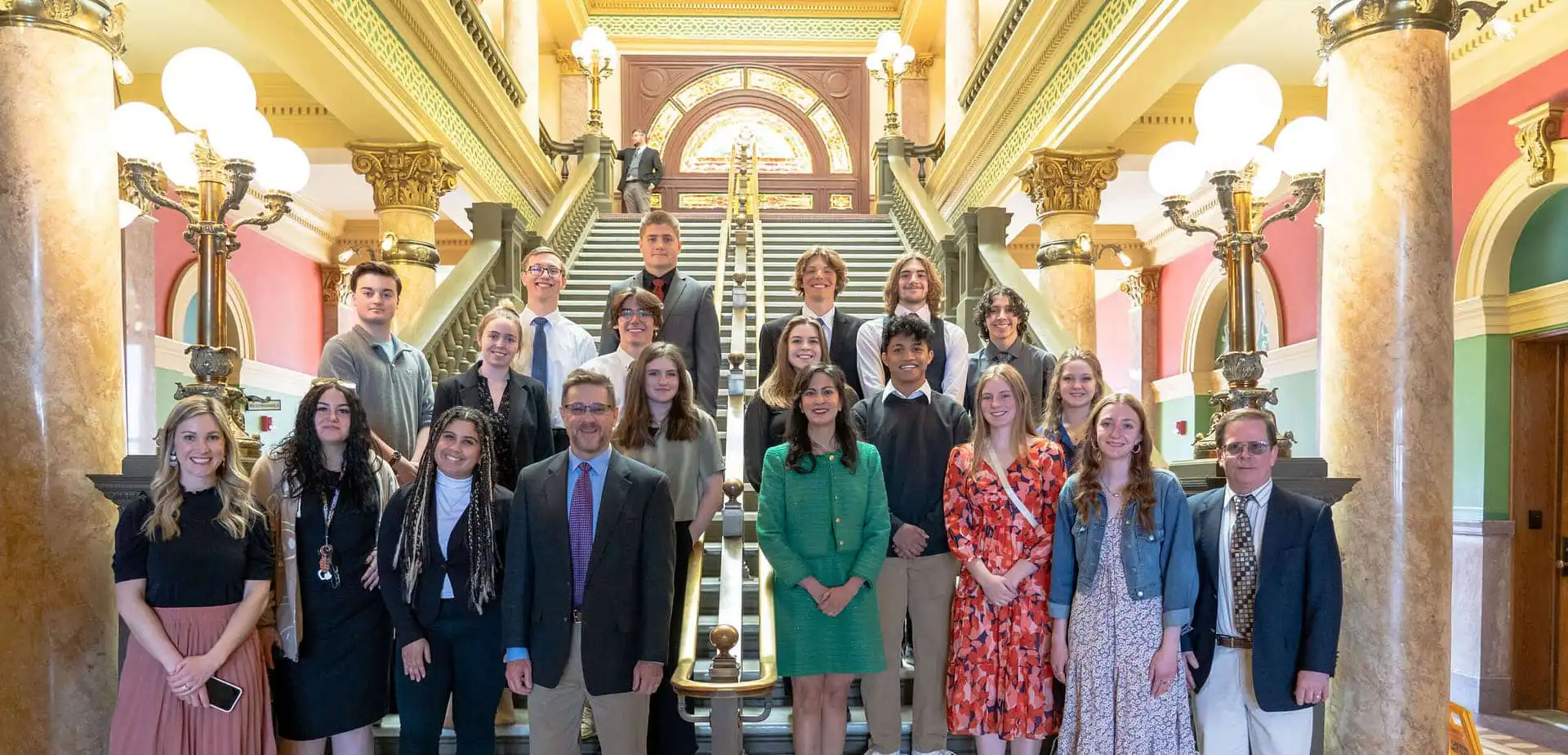 A group of people, including students from Stevensville High School and several adults, stand posing for a photo on a grand staircase inside a historic building with ornate columns, decorative lights, and colorful walls.