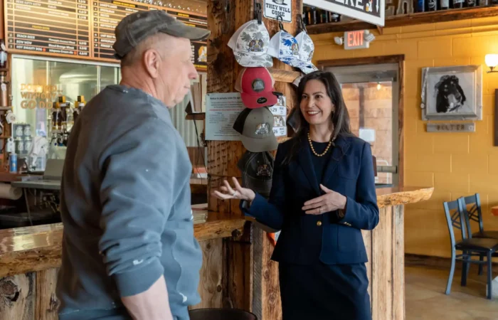 Secretary Christi Jacobsen notes Montana on pace for 3rd-straight year of record business registrations 3 A woman in a navy suit smiles and gestures while talking to a man in casual clothes and a cap inside a rustic bar or café, with hats displayed on the wall and a menu board in the background.