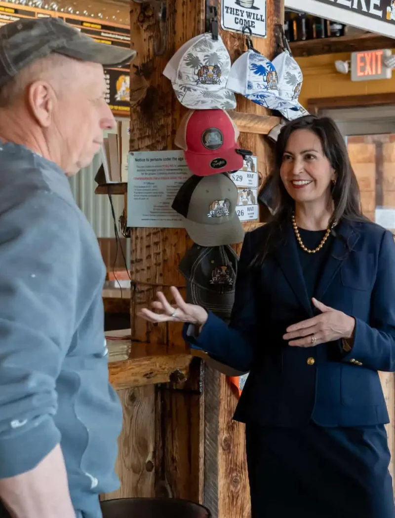 A woman in a navy suit smiles and gestures while talking to a man in casual clothes and a cap inside a rustic bar or café, with hats displayed on the wall and a menu board in the background.