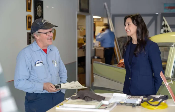 Secretary of State Christi Jacobsen visits White Sulphur Springs businesses, election officials, and students 5 A man in a blue work shirt and cap holds a certificate while talking to a woman in a navy blazer, possibly Secretary of State Christi Jacobsen. They stand by a counter with tools and papers, with a vintage car nearby in White Sulphur Springs.