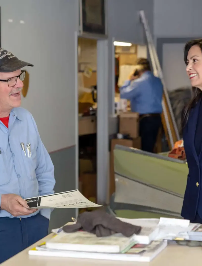 Secretary of State Christi Jacobsen visits White Sulphur Springs businesses, election officials, and students 5 A man in a blue work shirt and cap holds a certificate while talking to a woman in a navy blazer, possibly Secretary of State Christi Jacobsen. They stand by a counter with tools and papers, with a vintage car nearby in White Sulphur Springs.