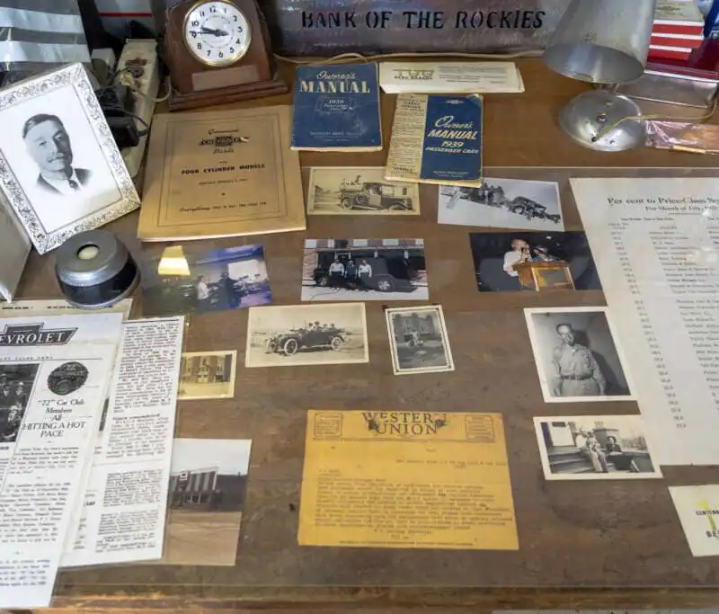 A wooden desk displays old photographs, documents, newspapers, a clock, vintage car manuals from Berg Garage, a framed black-and-white portrait, and a yellow Western Union telegram, creating a nostalgic Meagher County historical scene.