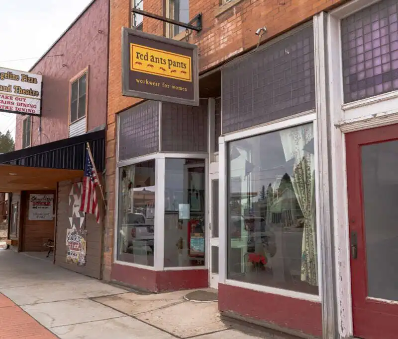 Street view of small-town brick storefronts, including Red Ants Pants—a Montana Business with a yellow sign and large windows—and a theater marquee displaying an American flag, all lining a quiet sidewalk.