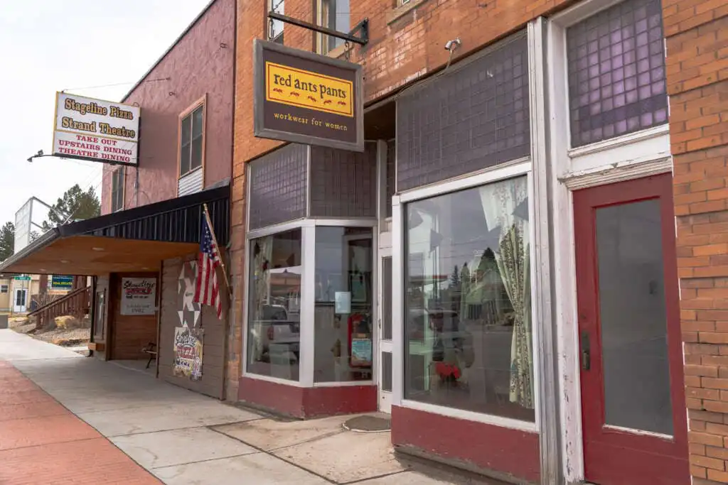 Street view of small-town brick storefronts, including Red Ants Pants—a Montana Business with a yellow sign and large windows—and a theater marquee displaying an American flag, all lining a quiet sidewalk.