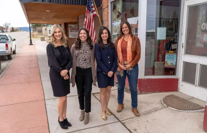 Montana Business Spotlight: ‘Fun to see what’s possible’ with Red Ants Pants 4 Four women stand smiling on a sidewalk in front of a storefront with an American flag. Three are dressed in business attire, and one in casual clothing with a scarf and boots. It appears to be a small town main street.