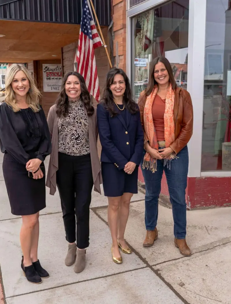 Four women stand smiling on a sidewalk in front of a storefront with an American flag. Three are dressed in business attire, and one in casual clothing with a scarf and boots. It appears to be a small town main street.