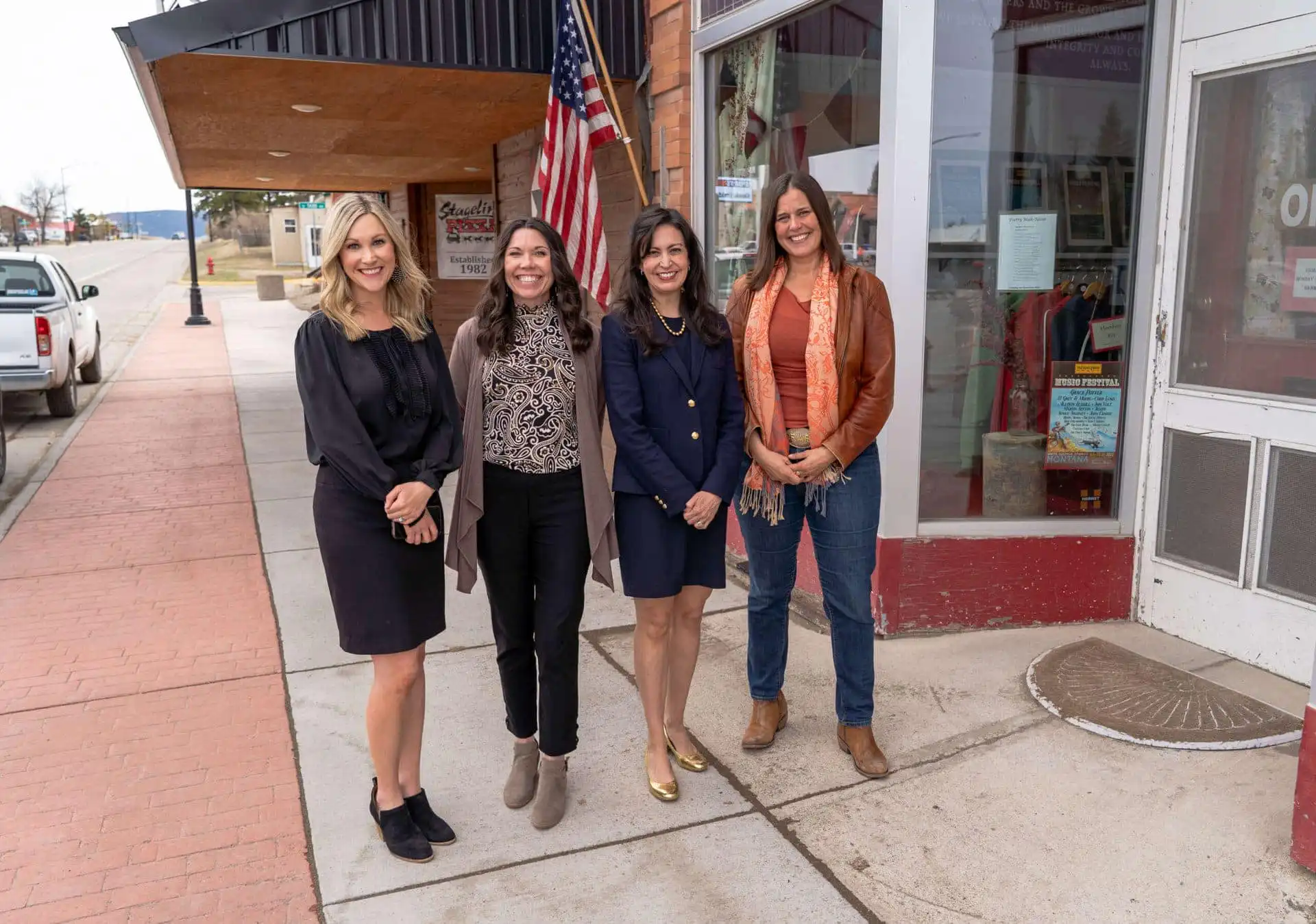 Four women stand smiling on a sidewalk in front of a storefront with an American flag. Three are dressed in business attire, and one in casual clothing with a scarf and boots. It appears to be a small town main street.