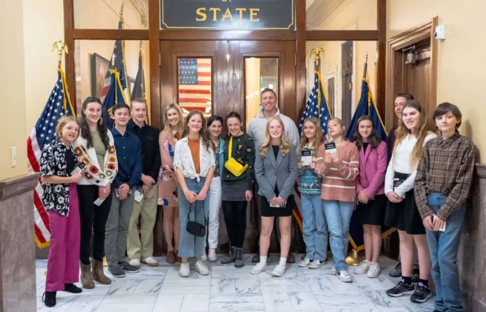 Red Lodge middle school students visit State Capitol, Secretary of State's Office 3 A group of teenagers from Red Lodge middle school and two adults stand together, smiling for a photo inside the State Capitol near marble floors, American flags, and a sign reading Secretary of State above wooden doors.