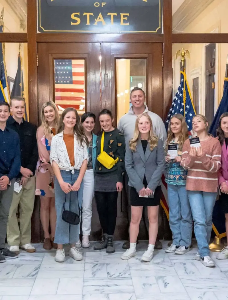A group of teenagers from Red Lodge middle school and two adults stand together, smiling for a photo inside the State Capitol near marble floors, American flags, and a sign reading Secretary of State above wooden doors.