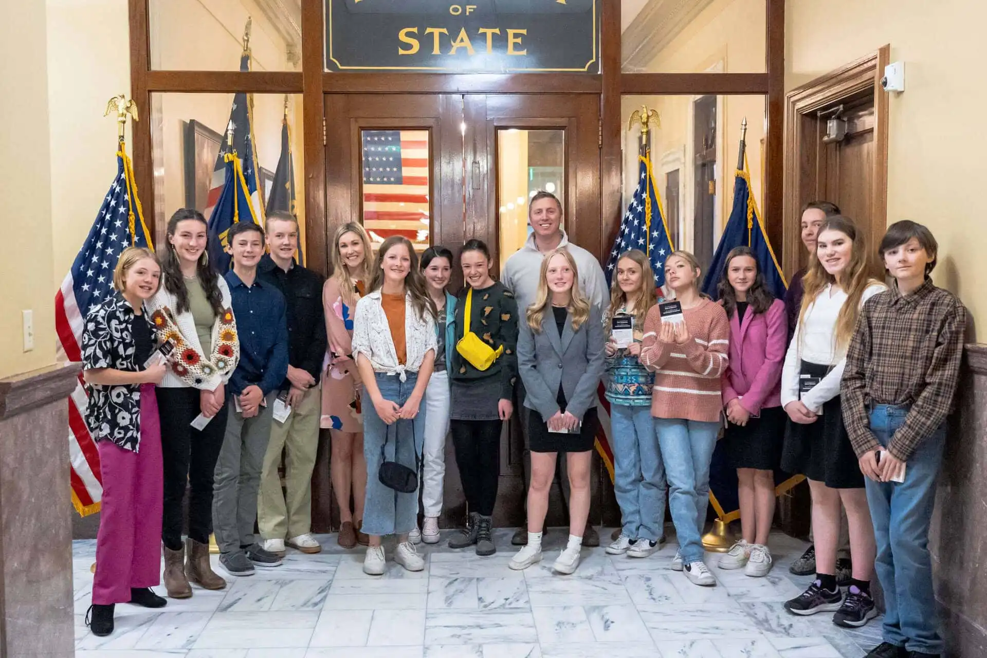 A group of teenagers from Red Lodge middle school and two adults stand together, smiling for a photo inside the State Capitol near marble floors, American flags, and a sign reading Secretary of State above wooden doors.