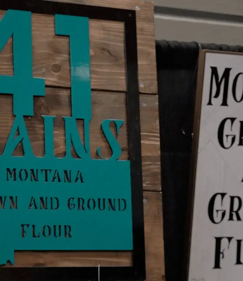 Wooden and turquoise sign reads “41 Grains Montana Grown and Ground Flour,” next to a white and black sign with similar text, both displayed at an indoor booth.