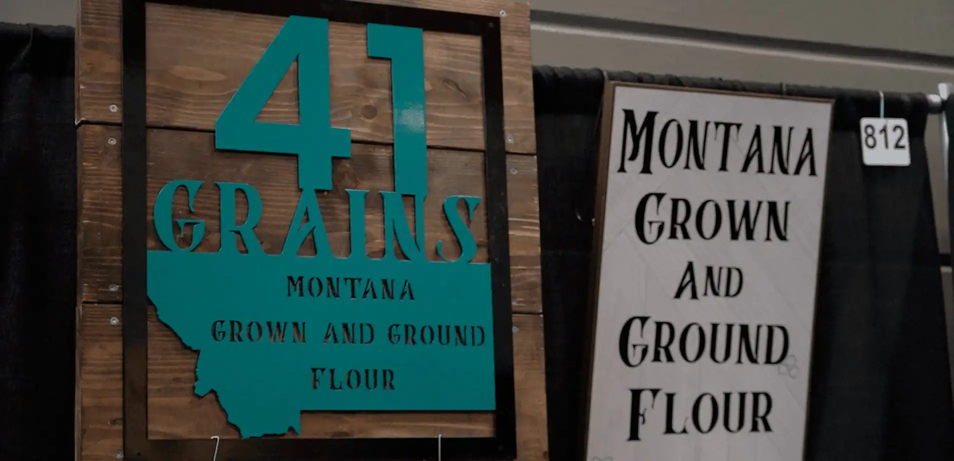 Wooden and turquoise sign reads “41 Grains Montana Grown and Ground Flour,” next to a white and black sign with similar text, both displayed at an indoor booth.