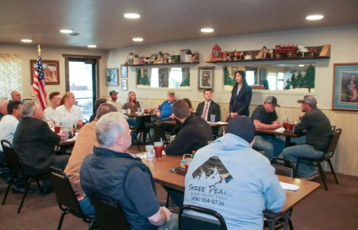 A group of people sit around tables in a restaurant-style room, listening to a woman speak about Bitterroot businesses. An American flag, framed pictures, and rustic decor are visible in the background.