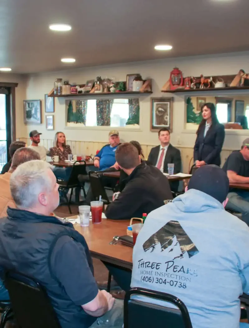 Secretary Christi Jacobsen visits with Bitterroot businesses, voters 4 A group of people sit around tables in a restaurant-style room, listening to a woman speak about Bitterroot businesses. An American flag, framed pictures, and rustic decor are visible in the background.
