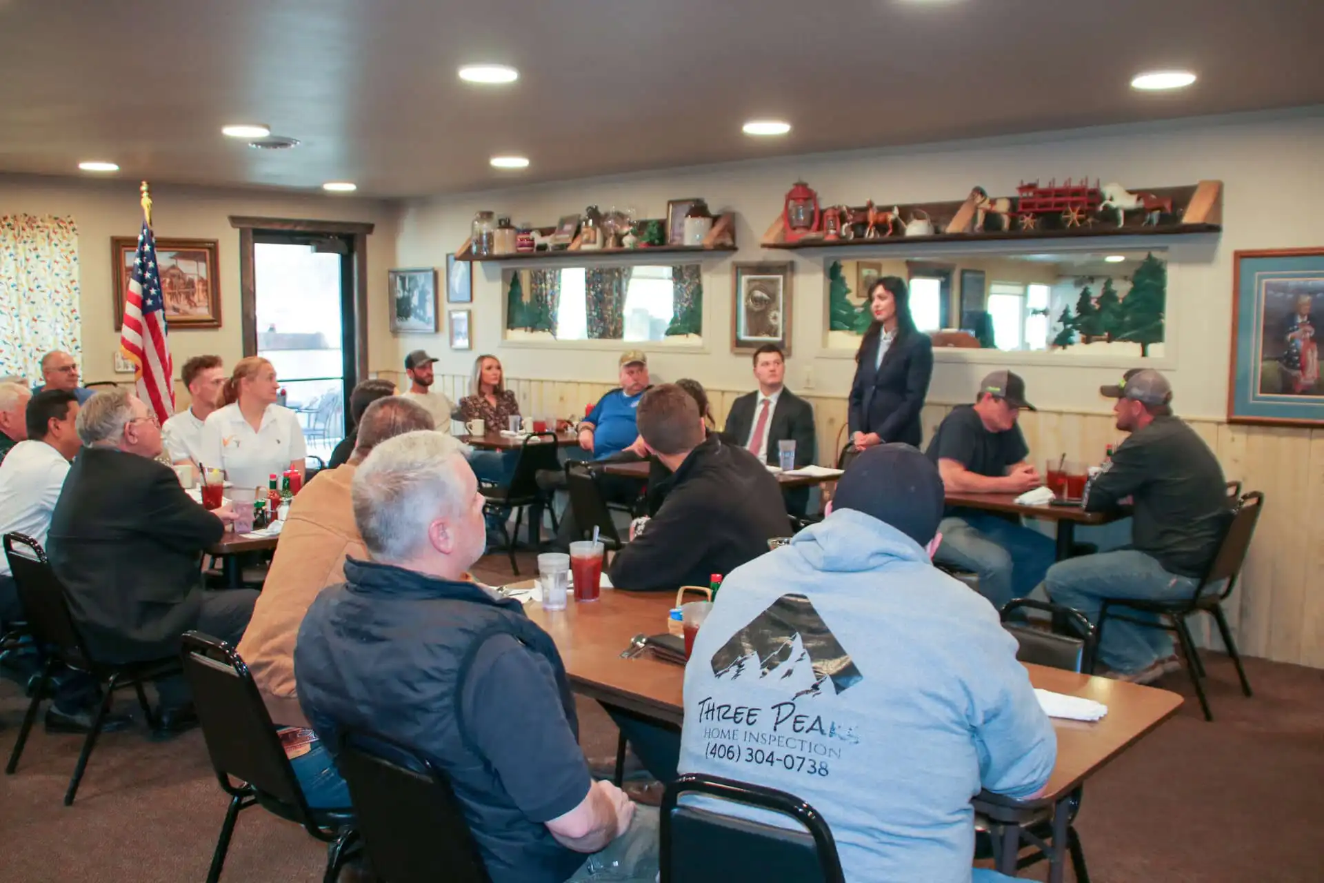 A group of people sit around tables in a restaurant-style room, listening to a woman speak about Bitterroot businesses. An American flag, framed pictures, and rustic decor are visible in the background.