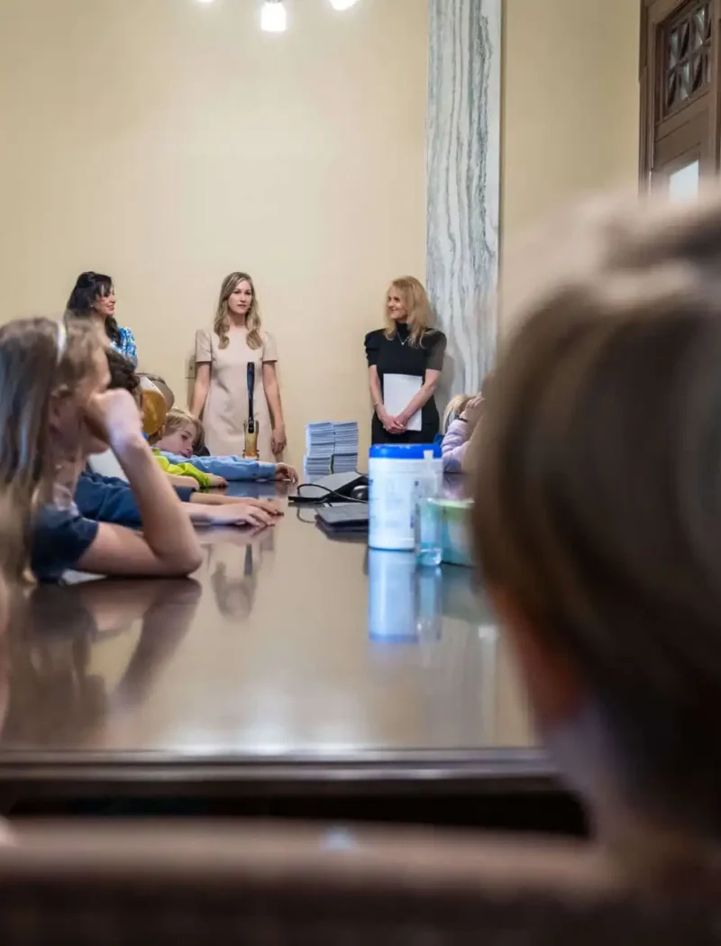 A group of children from Smith Elementary sit around a large table in a conference room at the Montana State Capitol, listening to Secretary Christi Jacobsen and two other women at the front. The atmosphere appears educational or formal.