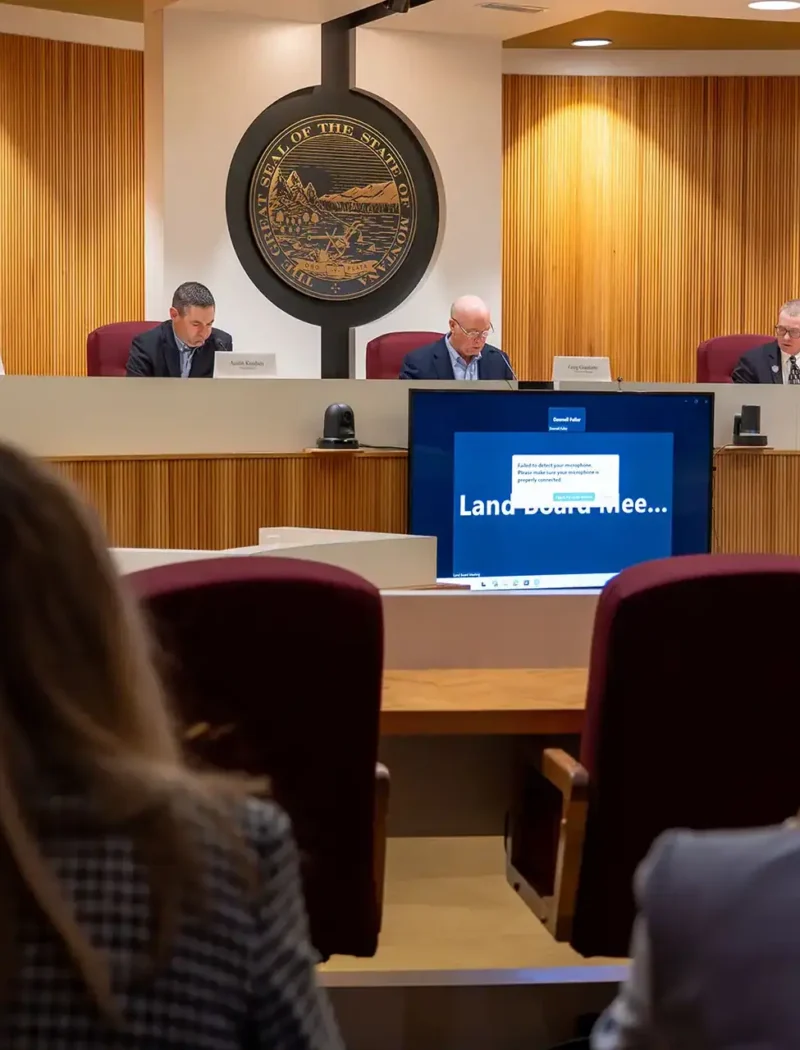 Five officials sit at a curved desk in a wood-paneled government meeting room, with a large seal on the wall and a screen displaying Land Use Committee. Two audience members are visible, as oil and gas projects and logging in Montana schools are discussed.