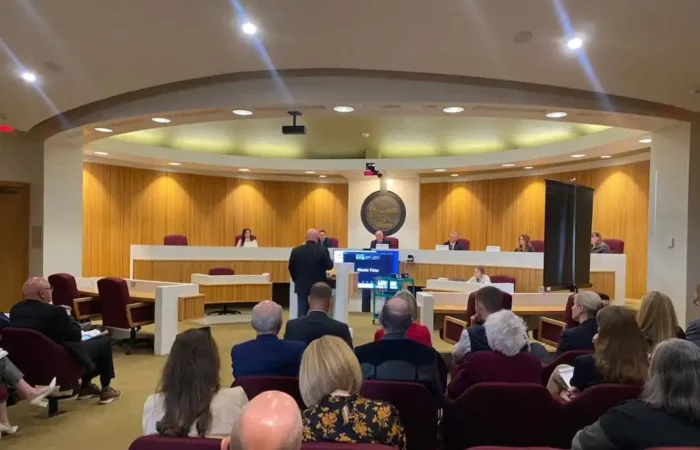 People sit in a Montana courtroom or council chamber, watching a speaker at the podium address Land Board Commissioners seated behind a long, curved bench. A monitor faces the audience, and the wood-paneled room has red chairs.