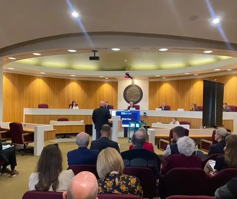 People sit in a Montana courtroom or council chamber, watching a speaker at the podium address Land Board Commissioners seated behind a long, curved bench. A monitor faces the audience, and the wood-paneled room has red chairs.