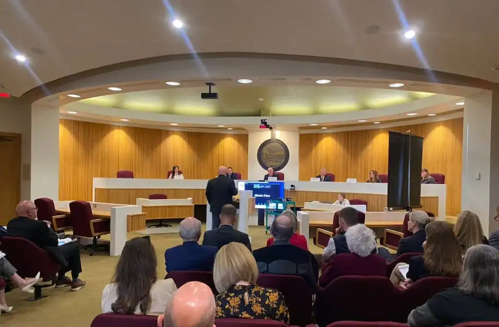 People sit in a Montana courtroom or council chamber, watching a speaker at the podium address Land Board Commissioners seated behind a long, curved bench. A monitor faces the audience, and the wood-paneled room has red chairs.