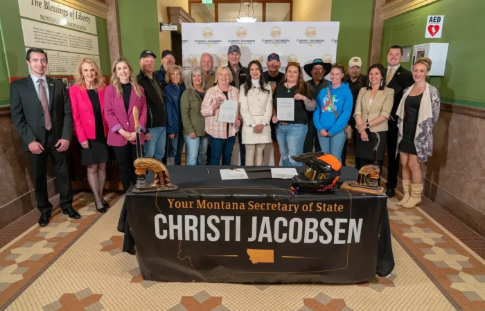Montana Snowmobile Association joins Secretary Christi Jacobsen to celebrate its 2025 legislative successes 1 A group poses indoors behind a table draped with a sign reading “Your Montana Secretary of State Christi Jacobsen.” Some hold a certificate; two gold helmets and a black helmet are displayed, celebrating Montana Snowmobile Association achievements.