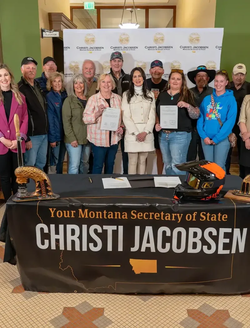 A group poses indoors behind a table draped with a sign reading “Your Montana Secretary of State Christi Jacobsen.” Some hold a certificate; two gold helmets and a black helmet are displayed, celebrating Montana Snowmobile Association achievements.