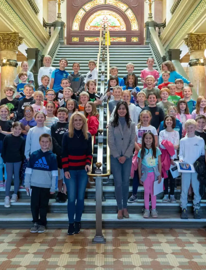 Montana City students and adults pose together on the steps of an ornate building with marble columns, a patterned tile floor, and a stained-glass window during their visit with the Secretary of State.